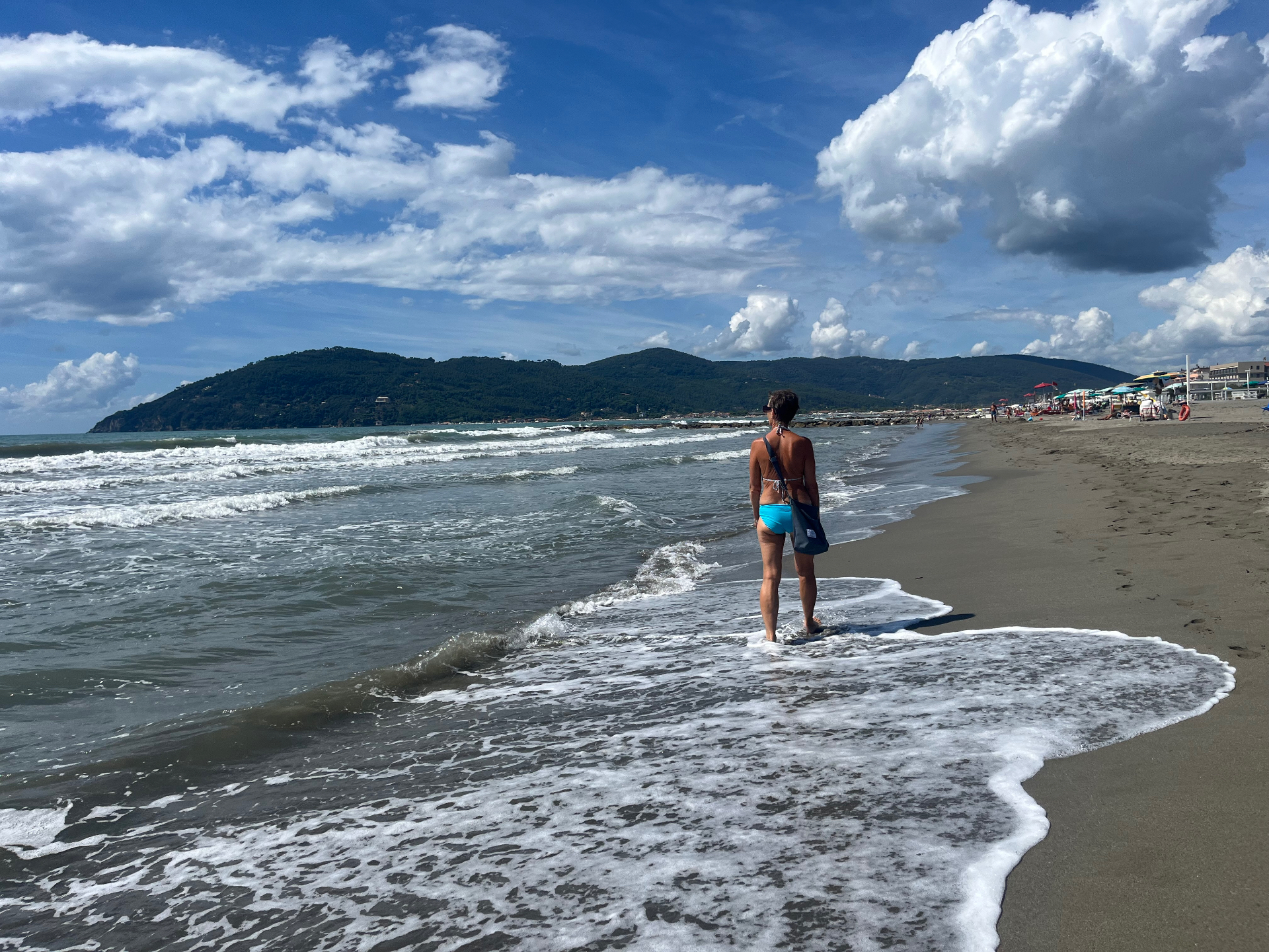 A walk along the water's edge on a sandy beach, with green hills and the shore lined with umbrellas beyond