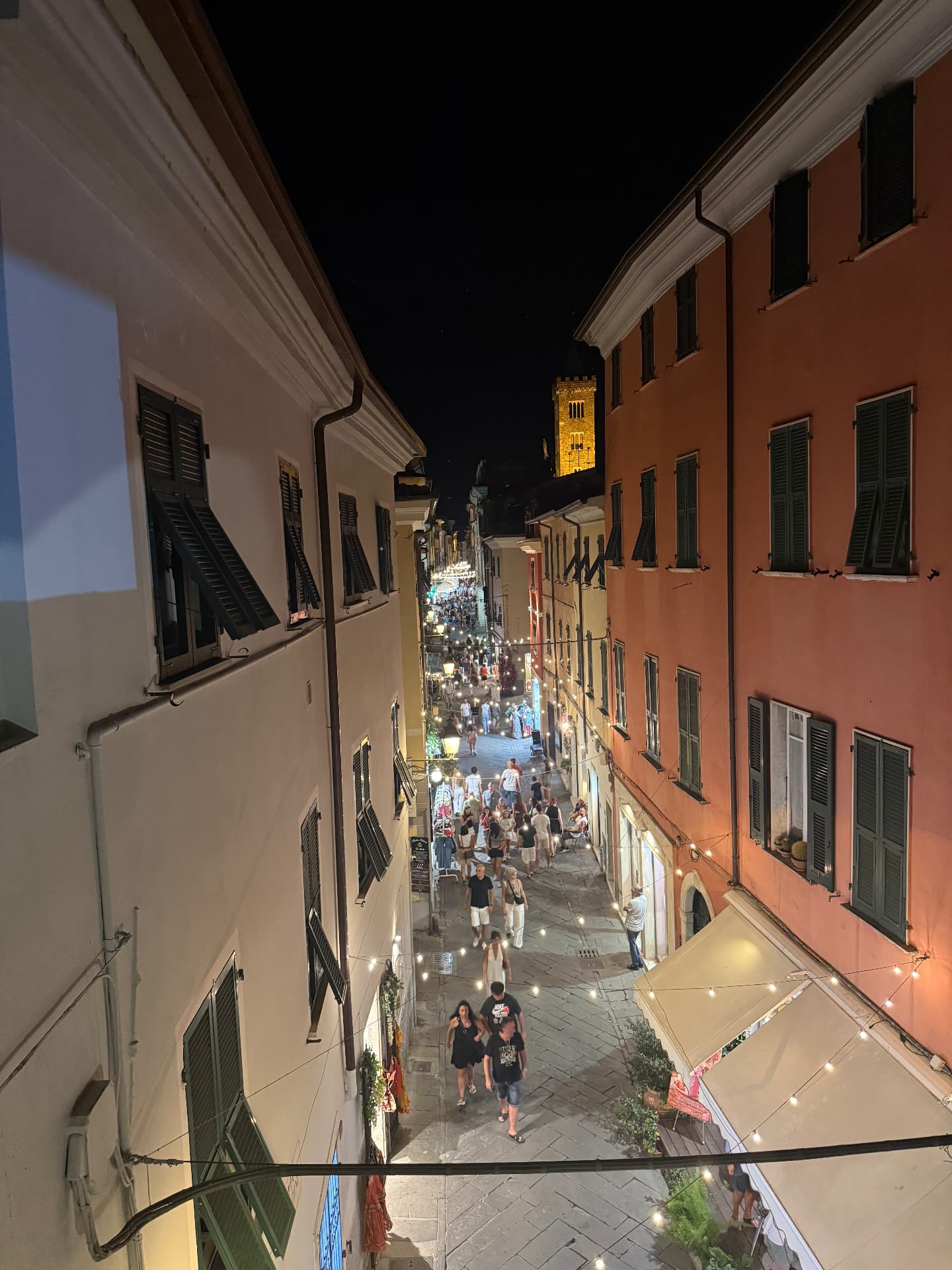 A lively pedestrian street at night with an illuminated bell tower in the distance