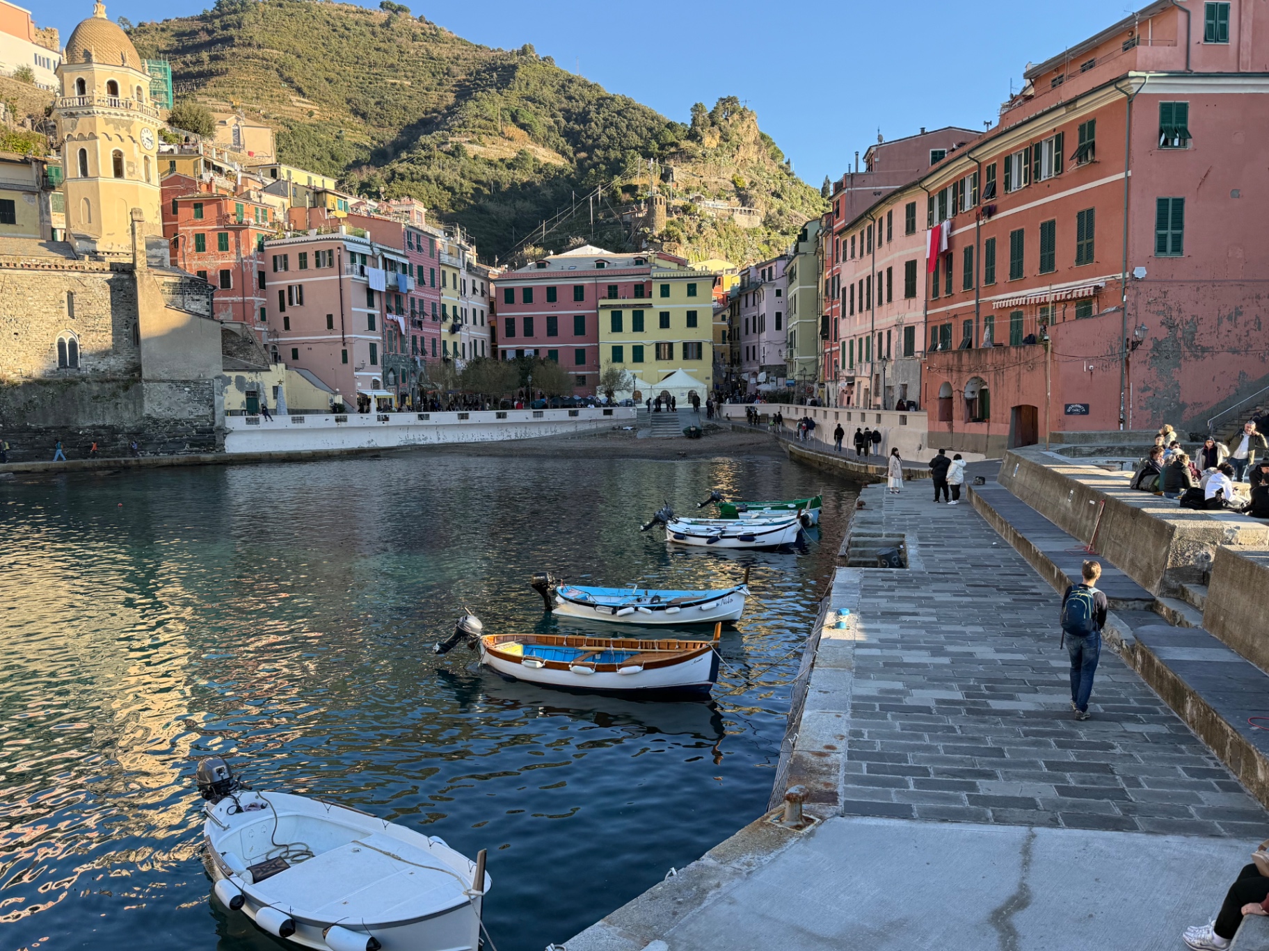 Small boats moored in a harbour on the Cinque Terre coast, below colourful houses and a stone church tower