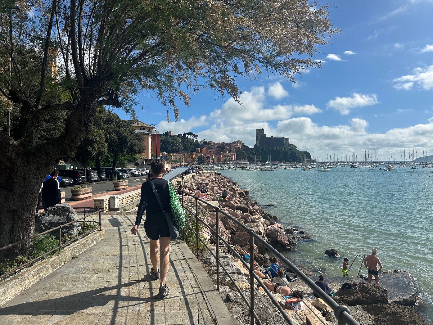Sailboats and sunbathers in the bay seen from the seafront promenade, a castle on the headland beyond